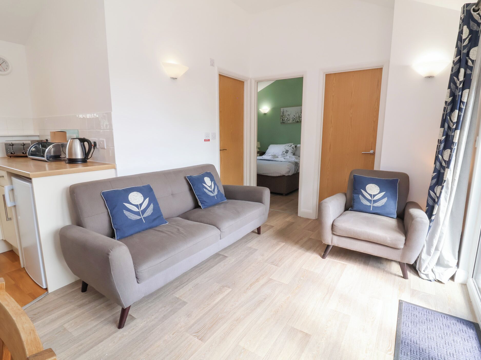 Un salon moderne avec un canapé et des fauteuils gris, un coin cuisine équipé d’une bouilloire, et une chambre à coucher visible à travers une porte ouverte.