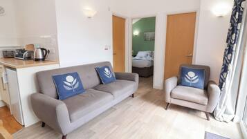 Un salon moderne avec un canapé et des fauteuils gris, un coin cuisine équipé d’une bouilloire, et une chambre à coucher visible à travers une porte ouverte.