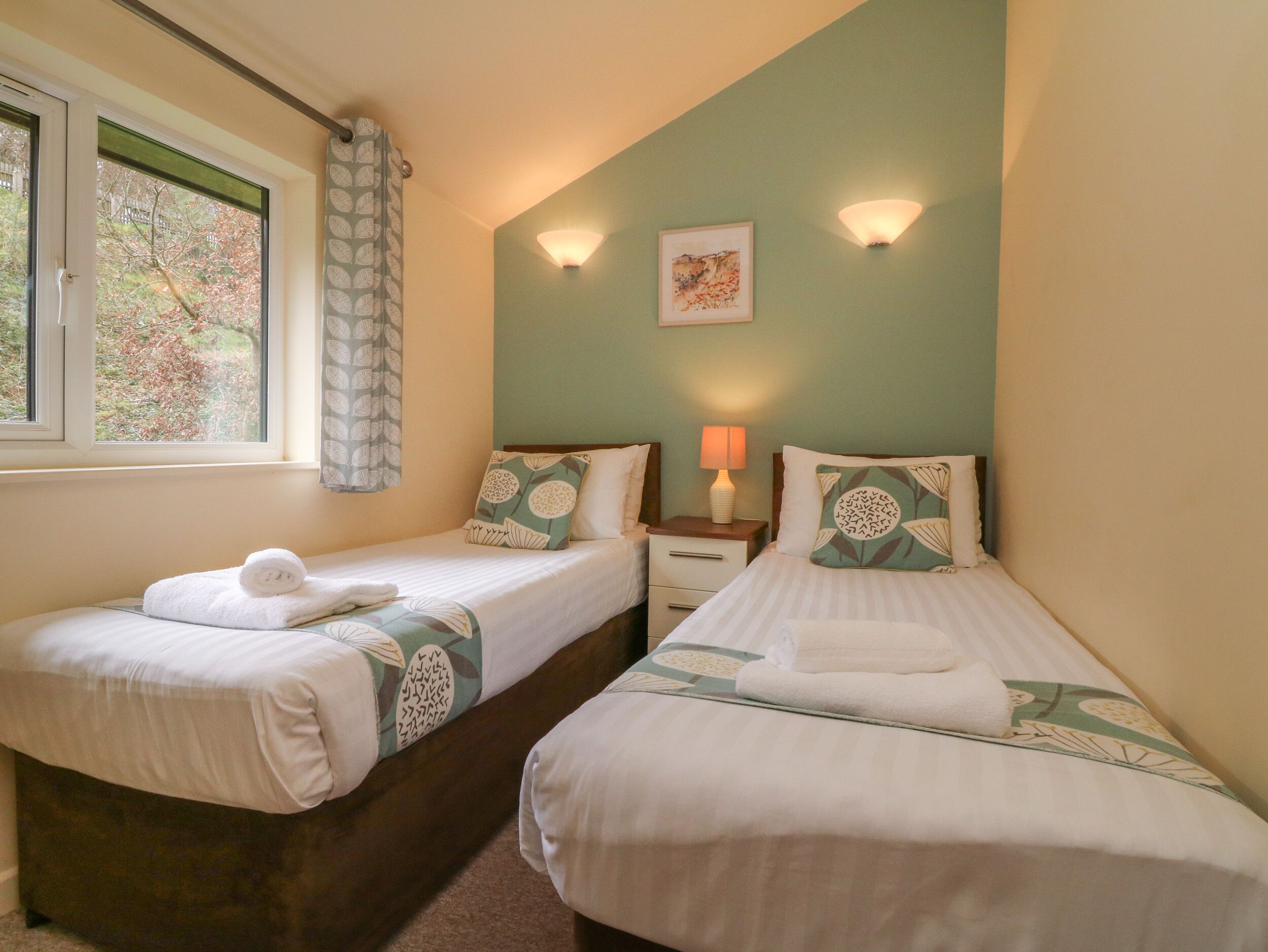 A bedroom with two beds, a window with a view of trees, and wall-mounted lamps.