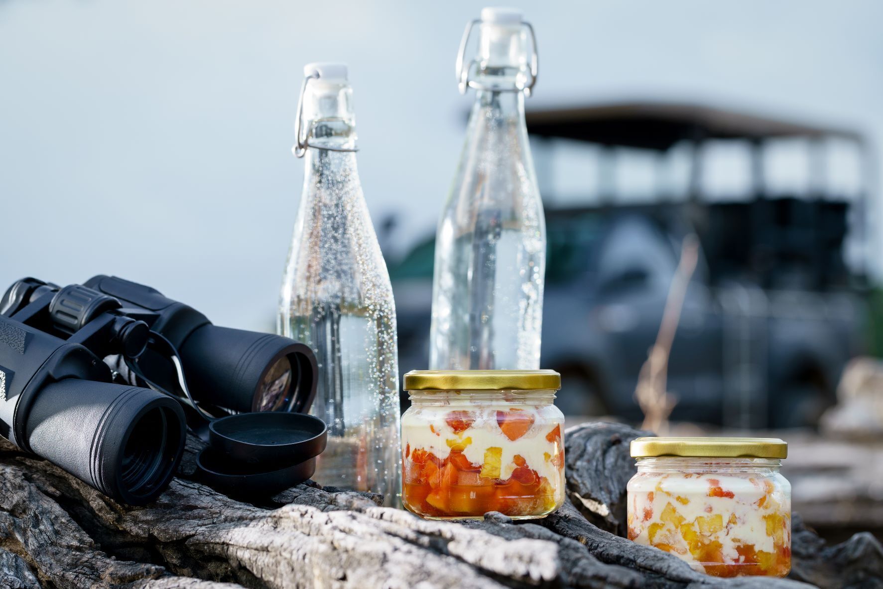 A pair of binoculars, two glass bottles, and two jars of preserved fruit on a wooden surface.