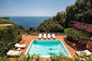 A swimming pool with lounge chairs, surrounded by greenery and overlooking the sea.