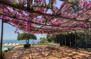 A covered outdoor area with a table and chairs, a pool, and a view of the sea.