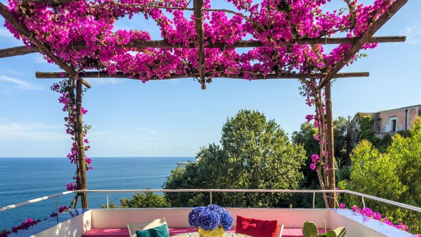 Une terrasse avec vue sur la mer, une table agrémentée d’un vase de fleurs et une pergola recouverte de fleurs roses.