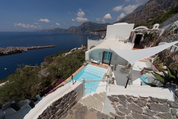 A modern villa with a pool overlooking the sea, stone walls, and a mountainous backdrop.