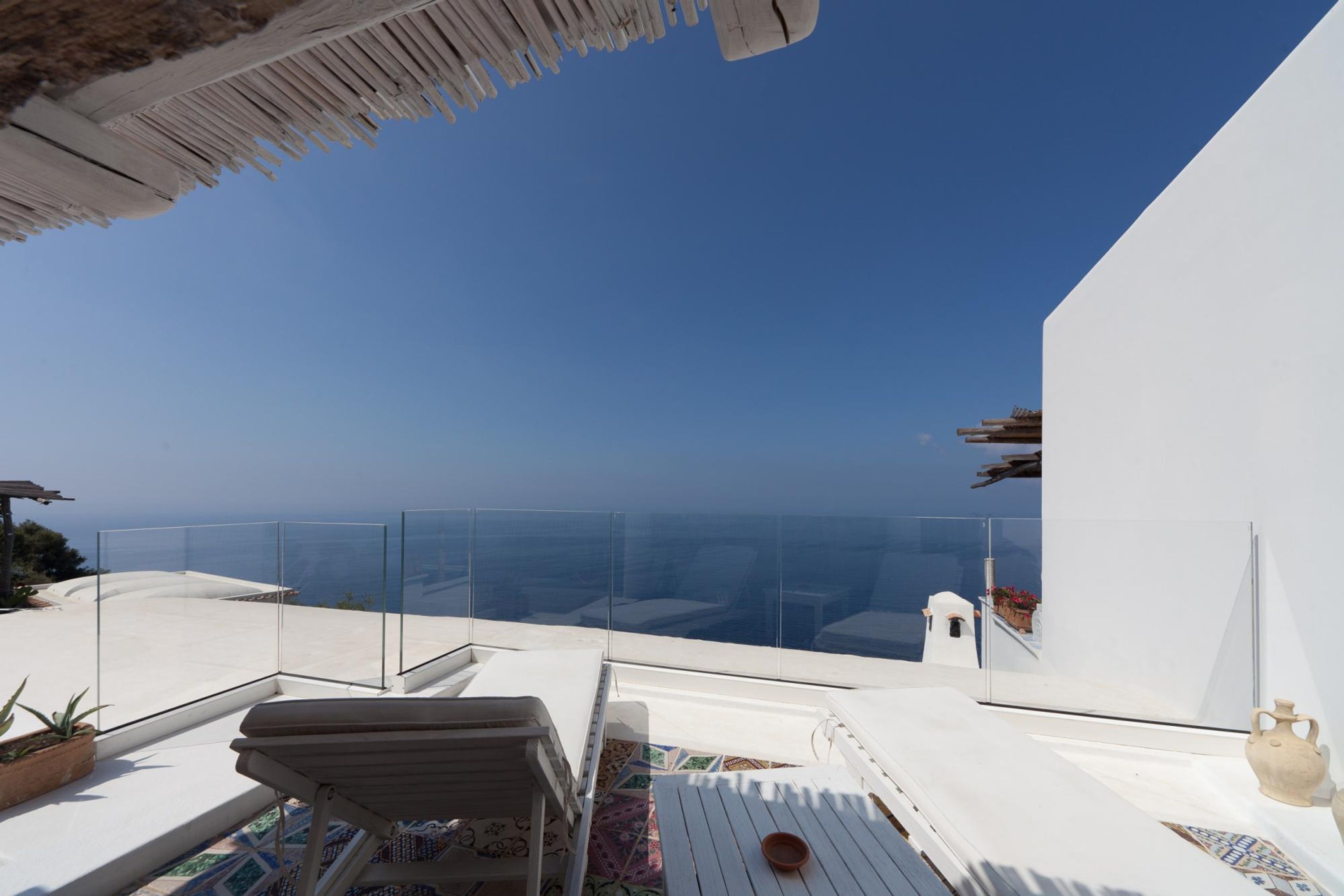 Eine Dachterrasse mit freiem Blick auf das Meer, weißen Möbeln und einem Glasgeländer.