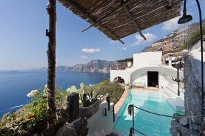 A pool with a view of the sea and mountains, a white building with an arched entrance, and a thatched roof structure.