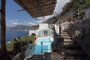 A pool area with stone steps leading down to a pool, surrounded by potted plants and a view of the sea.