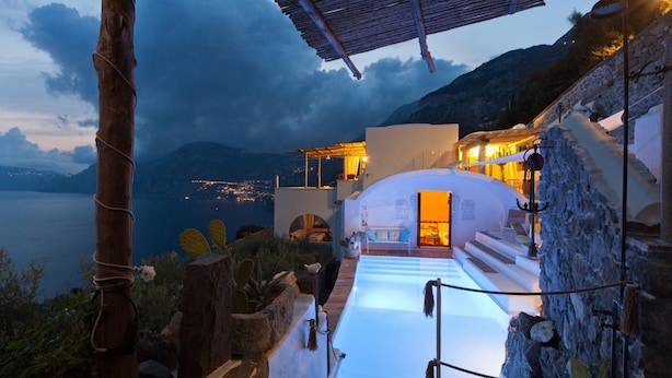 A modern villa with a glass infinity pool, stone walls, and a view of the sea.