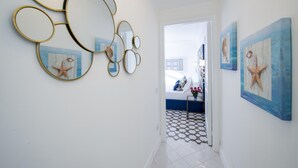 Un pasillo con suelo de baldosas, una puerta blanca que da a un dormitorio, y espejos y cuadros colgados en la pared.