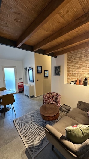 A living room with a sofa, a patterned armchair, a small round table, a red chair, and a rug. There is a brick wall with framed pictures and a window.