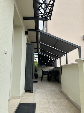 A covered outdoor seating area with a table and chairs, a potted plant, and a door leading to an interior space.