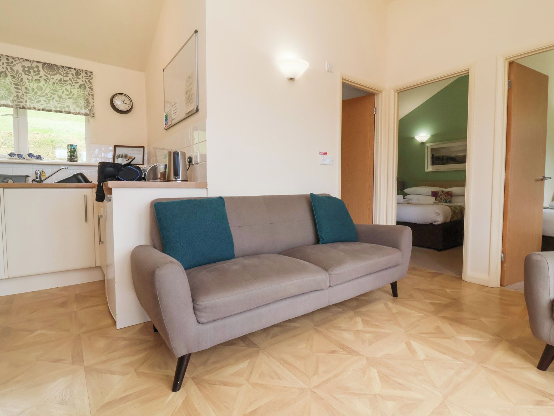 Un salón con un sofá gris, una zona de cocina con una tetera y otra tetera en la encimera, y un dormitorio con una cama y un cuadro en la pared.