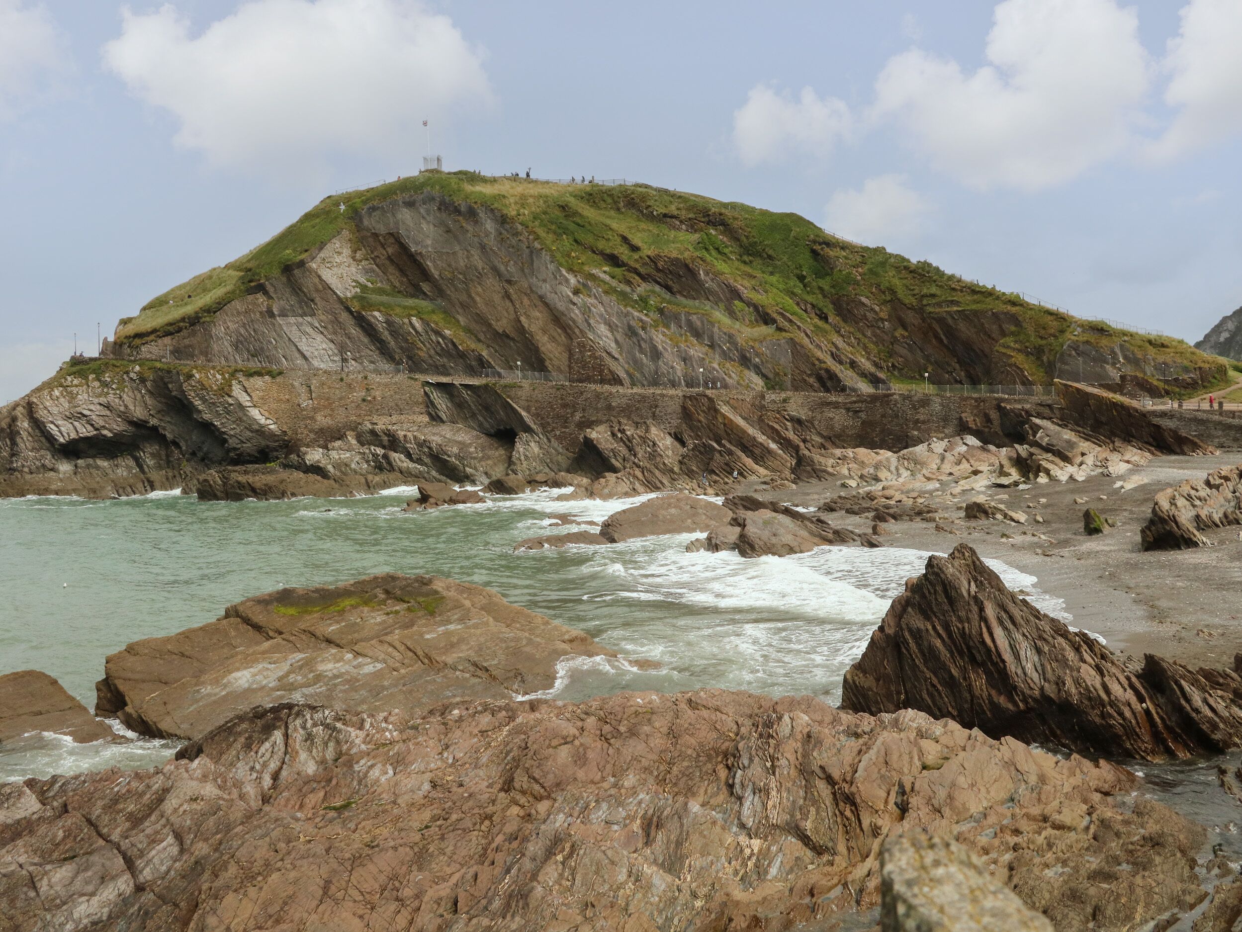 A rocky coastal landscape with a grassy hill and a bridge.