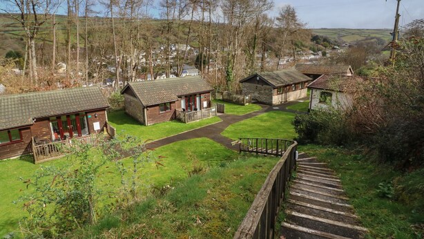 A rural setting with a small lodge featuring a porch and a pathway leading to a wooden bridge. - 7 Manleigh Park (Ilfracombe)