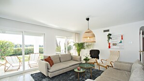 Ein modernes Wohnzimmer mit einer Couch, Sesseln, einem Couchtisch und einem großen Fenster mit Blick auf Grünflächen.