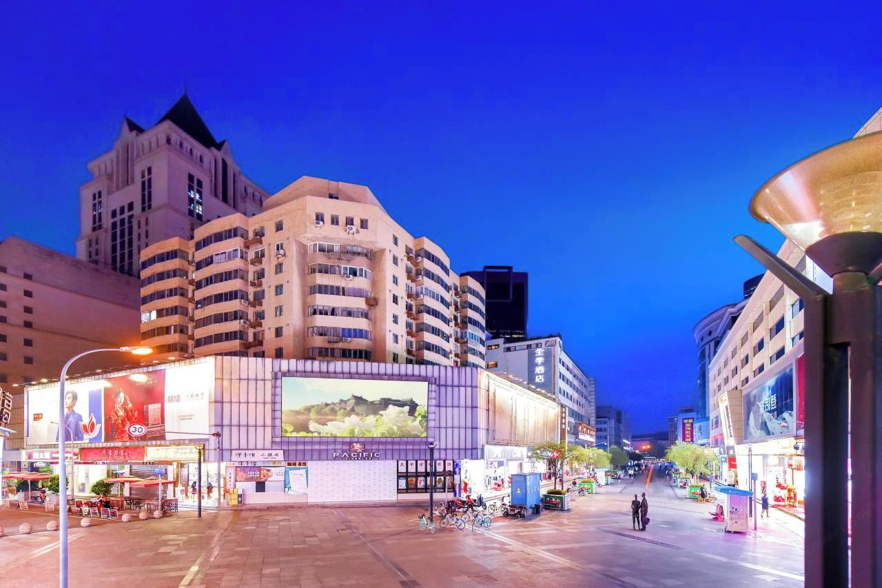 A cityscape at dusk with modern buildings, a large billboard, and a street with shops and a pedestrian area.