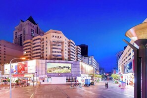 Une vue sur une ville au crépuscule, avec des bâtiments modernes, un grand panneau publicitaire et une rue bordée de boutiques et d’une zone piétonne.