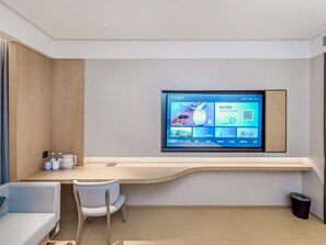 Une chambre d’hôtel équipée d’un téléviseur fixé au mur qui affiche l’interface de l’hôtel, d’un bureau en bois avec une chaise et d’une petite poubelle.