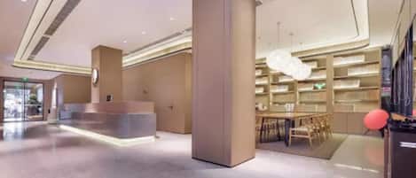 A modern hotel lobby with a reception desk, a large central column, and a dining area with a chandelier.