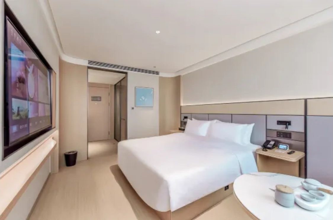 A modern hotel room with a large bed, bedside tables, a TV, and a small table with a cup and a book.