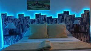 Un lit avec un drap blanc et deux oreillers, une table de chevet avec une lampe, et un mur orné d’une bande lumineuse bleue.
