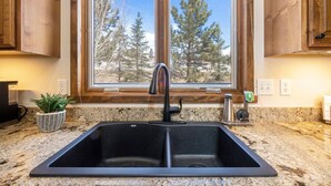 A kitchen with a double sink, wooden cabinets, a window with a view of trees, and a potted plant on the counter.