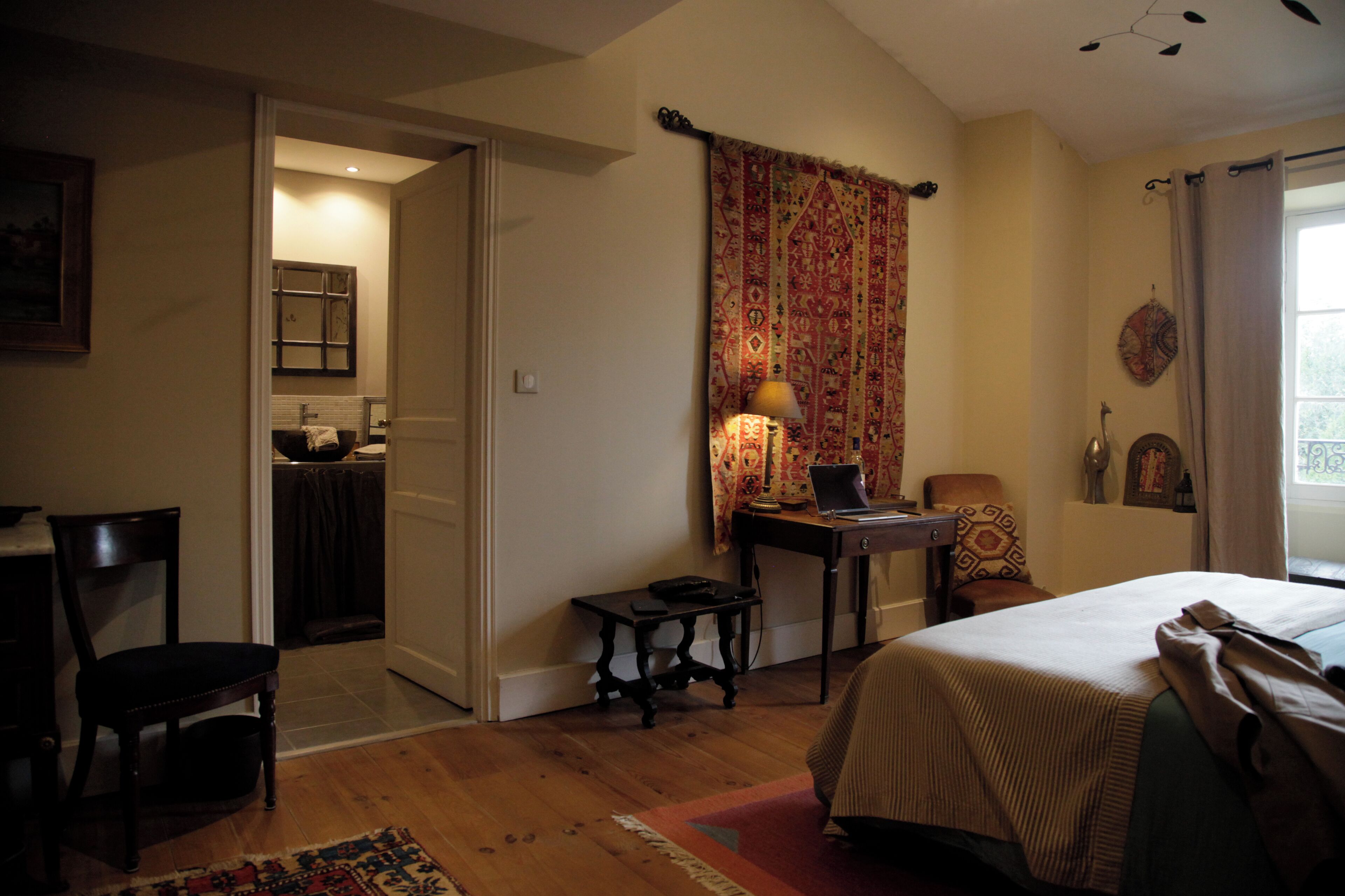 A bedroom with a bed, a desk with a laptop, a chair, a small table, and a window with curtains.