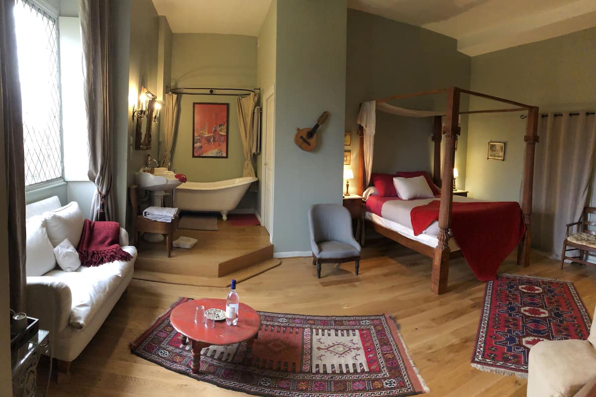 Une pièce avec un canapé blanc, une table rouge, un plancher en bois, un lit à baldaquin, une chaise, une petite table, une baignoire et une fenêtre avec des rideaux.