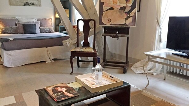 Een gezellige woonkamer met een bed, een houten stoel, een tafel met een fles en een pizzadoos, en een schilderij aan de muur.
