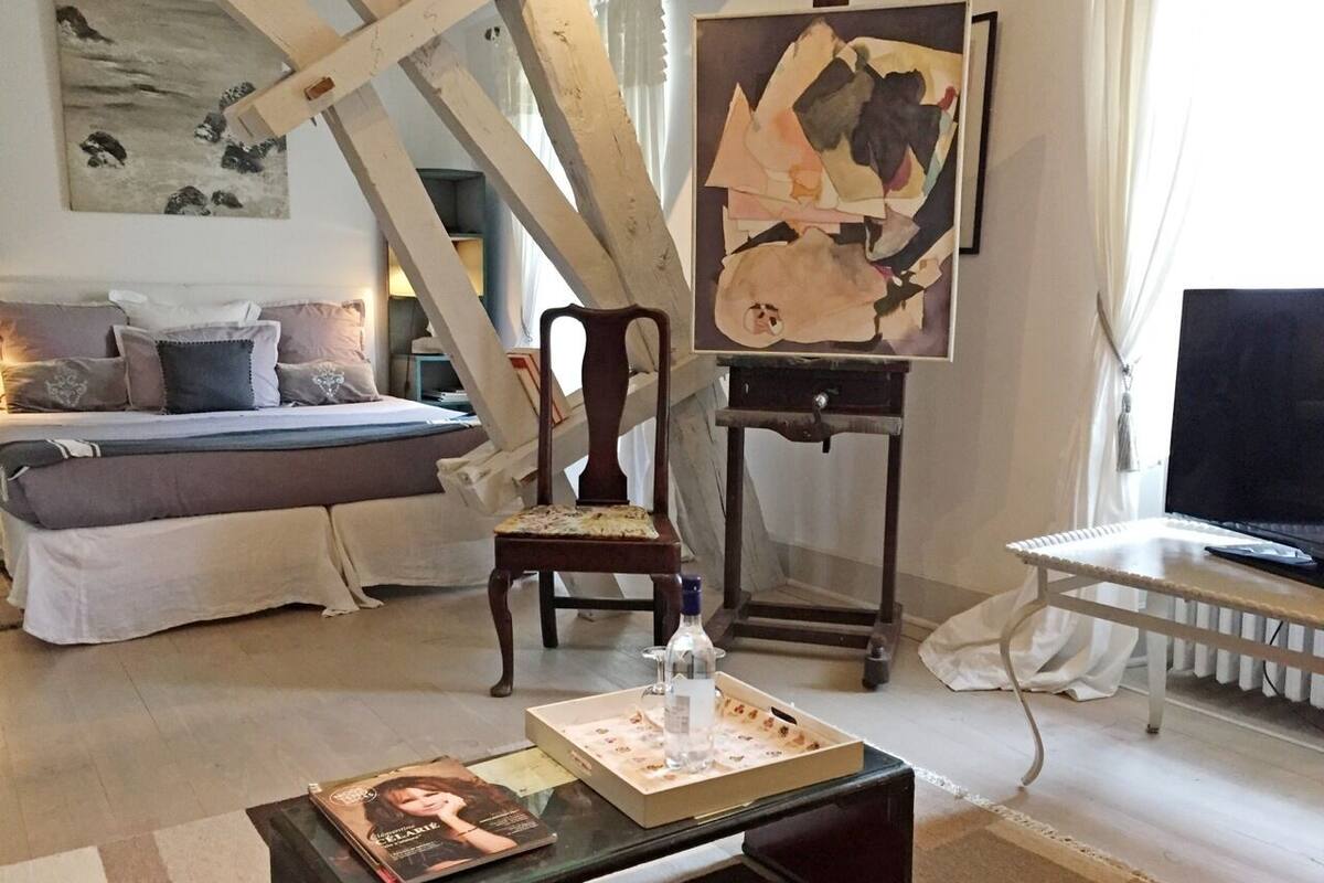 Un salon confortable avec un lit, une chaise en bois, une table sur laquelle se trouvent une bouteille et une boîte à pizza, et un tableau accroché au mur.