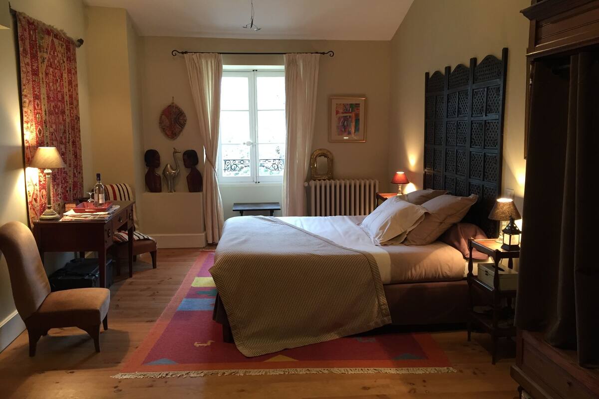 Une chambre à coucher comprenant un lit, des tables de chevet, une chaise, un bureau, une fenêtre avec des rideaux, un radiateur et une tête de lit fixée au mur.