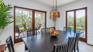 A dining area with a dark wooden table, six chairs, a woven fruit basket, and large windows with a view of a swimming pool and greenery.