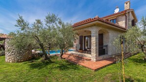 A stone house with a tiled roof, a pool, and a garden with olive trees.