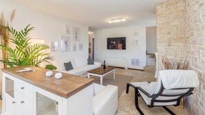 A modern living room with a white sofa, a wooden coffee table, and a TV mounted on the wall. There is a stone wall on the right and a plant on the left.