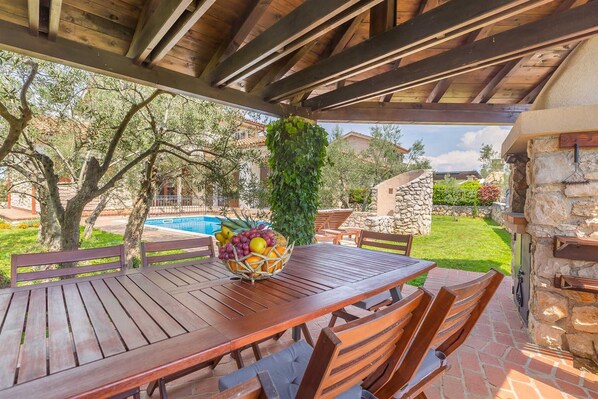 An outdoor wooden table with chairs, a fruit basket, and a stone wall with a grill.