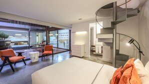 Et moderne hotelværelse med seng, orange stole, et lille bord, en trappe og udsigt over hotellets indre.