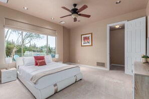 A spacious bedroom with a large bed, a ceiling fan, a window with a view of cacti, and a door leading to another room. - Desert Oasis ★Pool-Spa-Disc Golf-Ping Pong-Poker Table-Pool Table★ (Scottsdale)