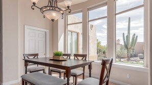 A dining area with a wooden table and chairs, a chandelier, and large windows with a view of cacti and a building outside. - Desert Oasis ★Pool-Spa-Disc Golf-Ping Pong-Poker Table-Pool Table★ (Scottsdale)