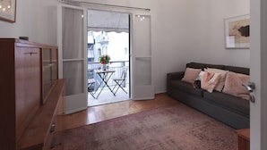 Ein Wohnzimmer mit grauem Sofa, einem Holzschrank, einer Chandelüster und einem Balkon mit Tisch und Stühlen.