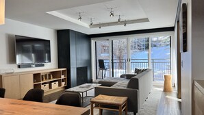 A modern living room with a large TV, a sofa, a dining table, and a view of a snowy landscape through large windows. - monarch chalet aspen condo (Aspen)