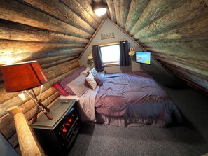 Ein gemütliches Schlafzimmer im Dachgeschoss mit schrägen Wänden, einem Bett mit Kopfteil, einem Nachttisch, einer Lampe, einem Fernseher und einem Fenster mit Vorhängen.