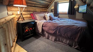 A cozy attic bedroom with a wooden ceiling, a bed with a purple bedspread, a red lamp, a fireplace, and a window with curtains. - Tiny Cabin in Huron National Forest! (Luzerne)