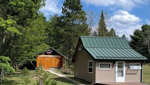 A small cabin with a green roof, a wooden shed, and a signpost in a grassy area with trees in the background. - Tiny Cabin in Huron National Forest! (Luzerne)