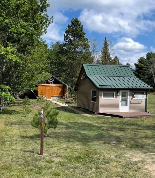 A small cabin with a green roof, a wooden shed, and a signpost in a grassy area with trees in the background. - Tiny Cabin in Huron National Forest! (Luzerne)