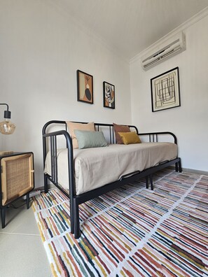 A bedroom with a black metal bed frame, a beige mattress, and colorful striped rug. There are two framed abstract paintings on the wall and a small wooden cabinet with a woven front. - Modern family home (Faro)