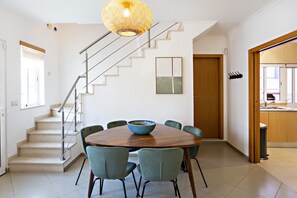 Un coin repas avec une table ronde en bois, six chaises, un saladier, un escalier et une cuisine visible à travers une porte ouverte.