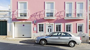 A pink building with three white-framed windows and balconies, a white garage door, and a parked silver car.