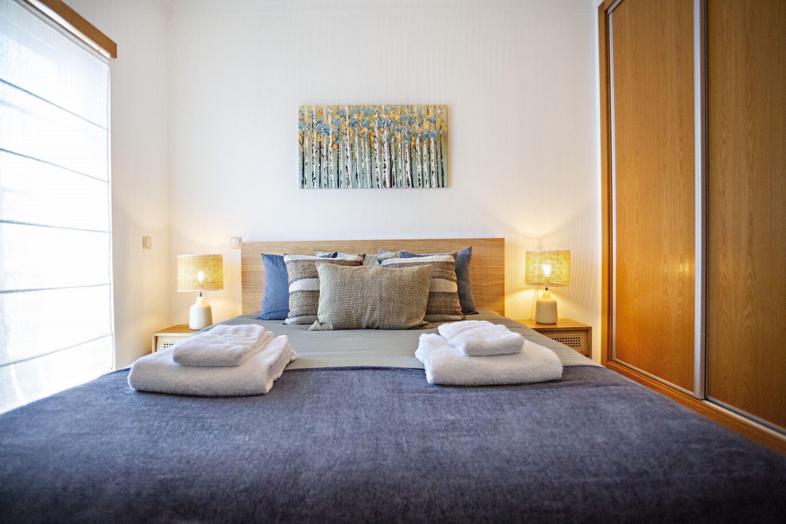 A neatly made bed with two towels, a painting on the wall, and two bedside lamps.