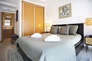 Une chambre à coucher bien rangée, avec un grand lit, une armoire en bois, une table de chevet avec une lampe et une œuvre d’art accrochée au mur.