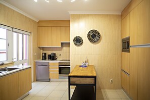 A compact kitchen with wooden cabinets, a dishwasher, an oven, and a microwave. There is a window with blinds, a wooden table, and two decorative plates on the wall. - Modern family home  (Faro)